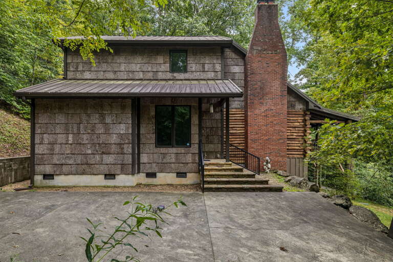 Rustic mountain cabin with distinctive brick chimney and charming shake siding, surrounded by lush greenery in a serene wooded setting.