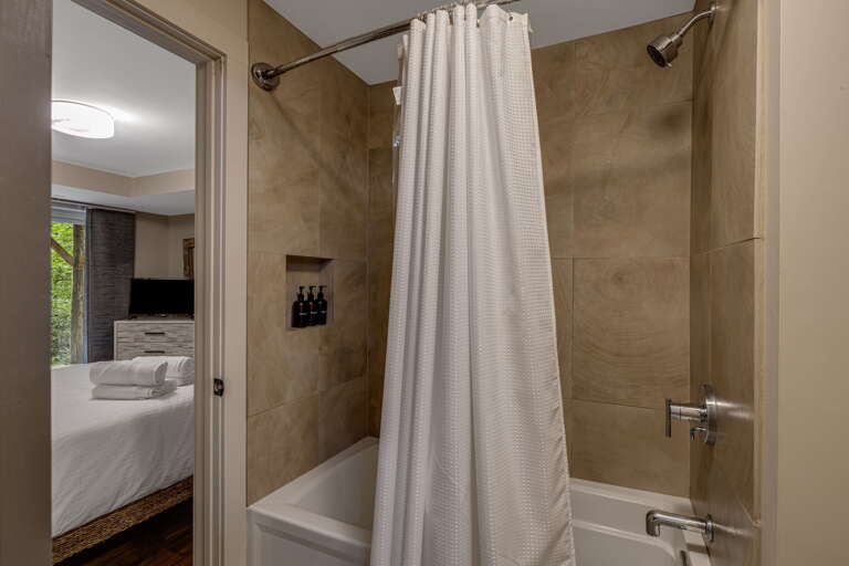 Fresh and serene, your private bathroom features a full tub-shower combo with waffle-weave curtain, warm tile surround, and soft lighting. Beyond the doorway, your bedroom awaits with plush towels ready for you.