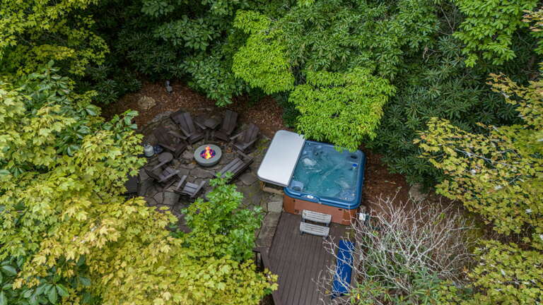 Aerial view reveals a private outdoor retreat featuring a hot tub and fire pit seating area nestled within lush, mature landscaping—perfect for quiet evenings under the trees.