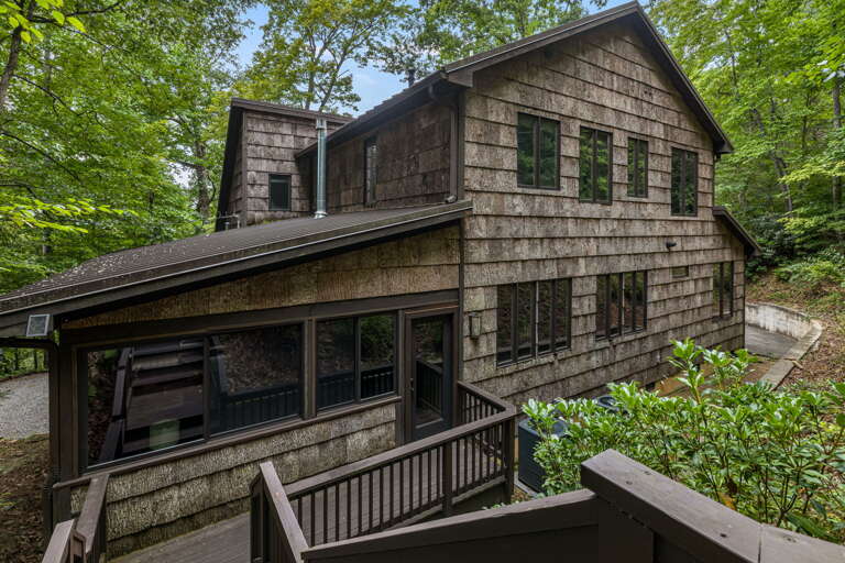 A two-story cedar-shingled retreat nestled among lush summer trees, with a welcoming covered deck framed by greenery and dappled forest light.