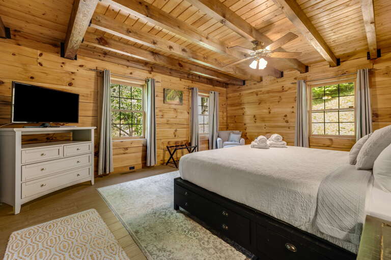 Relax in your serene bedroom with soaring wooden beams, cozy linens, and natural light streaming through windows overlooking lush greenery.
