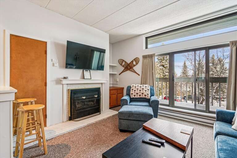 Interior Living Space With Sofa, Armchair, Television, And Fireplace, Opening To Forested View Through Large Window