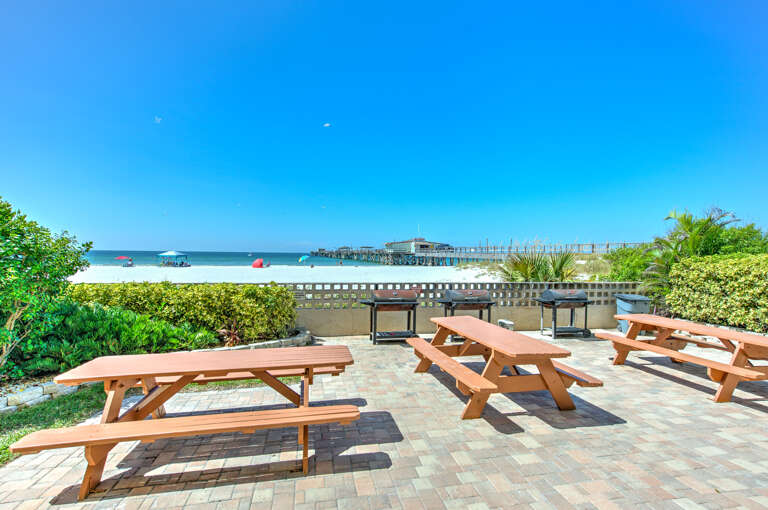 Beachfront dining area with wooden picnic tables overlooking pristine white sand and turquoise waters, perfect for outdoor meals.