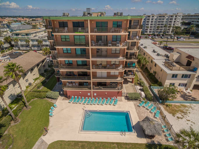 Aerial view of the beachfront property featuring a sparkling pool surrounded by turquoise lounge chairs and lush landscaping in a coastal neighborhood.