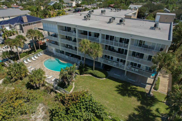 Aerial view of a three-story vacation rental property featuring a sparkling pool surrounded by tropical landscaping and palm trees.