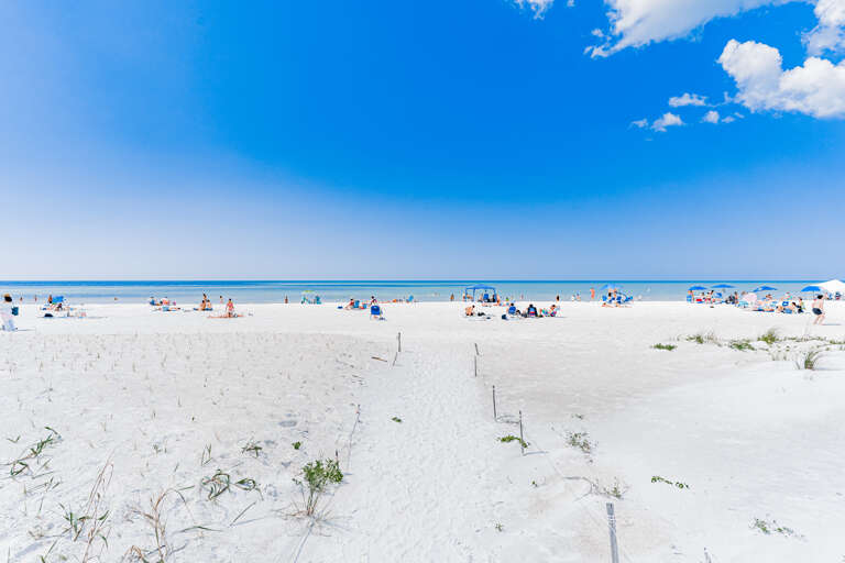 Pristine white sand beach stretches endlessly under brilliant blue skies, with gentle waves and scattered beachgoers enjoying the perfect coastal setting.