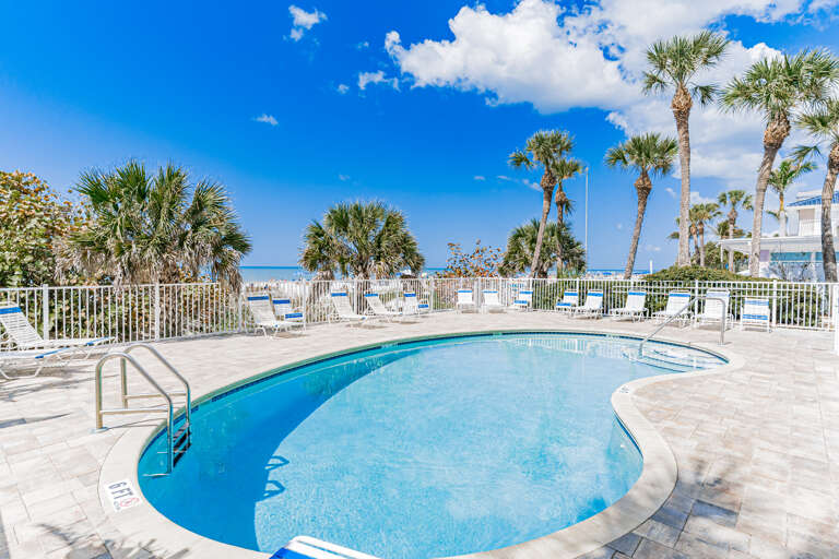 Beachfront pool area featuring crystal-clear water, palm trees, and direct beach access under brilliant blue skies.