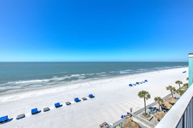 Pristine white sand beach stretches along crystal-clear waters under brilliant blue skies with rental umbrellas and palm trees dotting the shoreline.