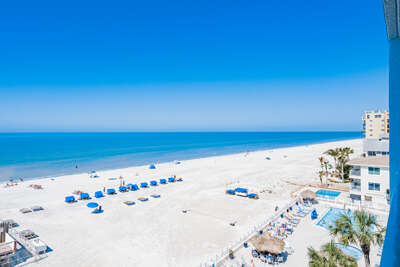 Pristine white sand beach stretches along crystal blue waters, with palm trees and beachfront buildings completing the coastal scene.