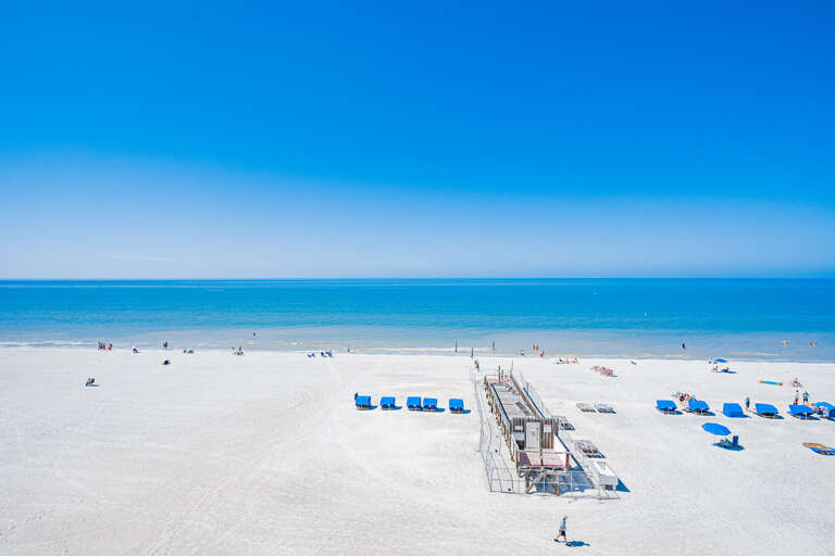 Pristine white sand beach stretches along crystal-clear turquoise waters under brilliant blue skies, with beach chairs and boardwalk access for guests.