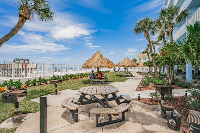 Tropical beachfront property featuring thatched-roof cabanas, picnic tables, and lush palm landscaping with direct beach access.