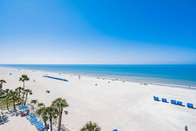 Pristine white sand beach stretches along turquoise waters under clear blue skies, with palm trees and beach chairs dotting the shoreline.