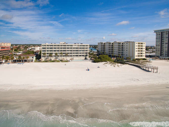 Aerial view of beachfront property buildings along pristine sandy shoreline with clear blue sky and gentle waves.