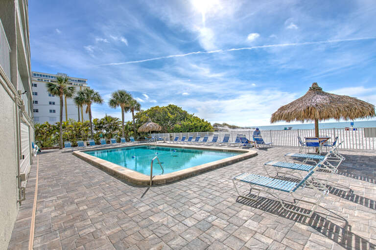 Beachfront pool area with tiki umbrella, lounge chairs, and palm trees under bright blue skies near the white sand beach.