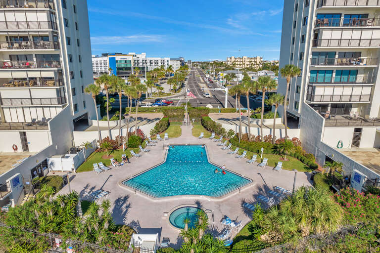Aerial view of a modern resort complex with swimming pool and landscaped gardens, surrounded by mid-rise buildings and palm trees.