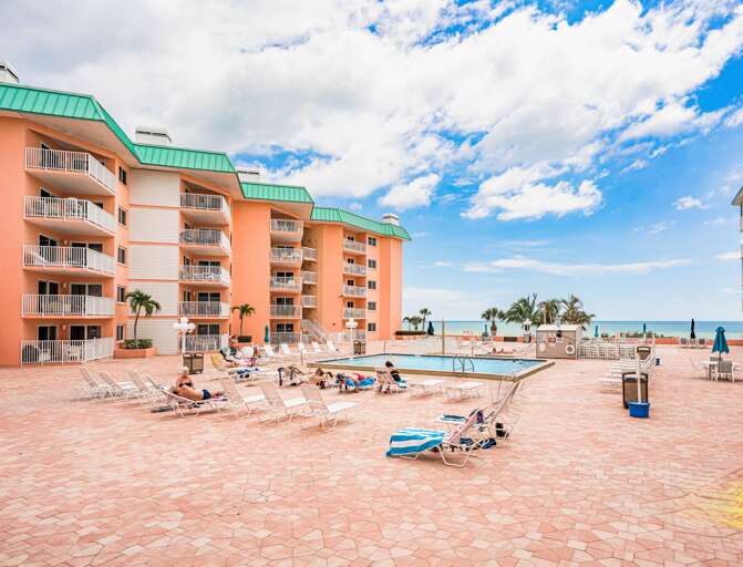 Beachfront resort property with coral-colored buildings, green roofs, pool area, and direct beach access under sunny skies.