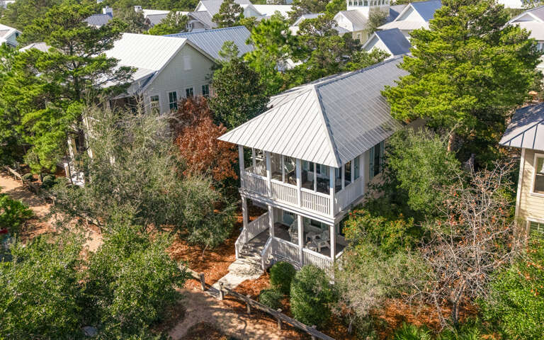 Aerial view of a charming coastal home with wraparound porches, nestled among mature trees in a peaceful neighborhood setting.