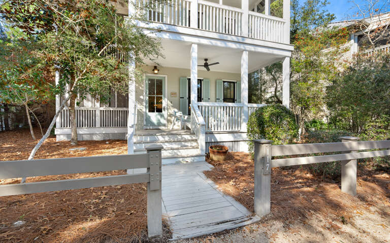 Charming cottage features welcoming front porch with ceiling fan and classic white railings, nestled among mature trees in peaceful residential setting.