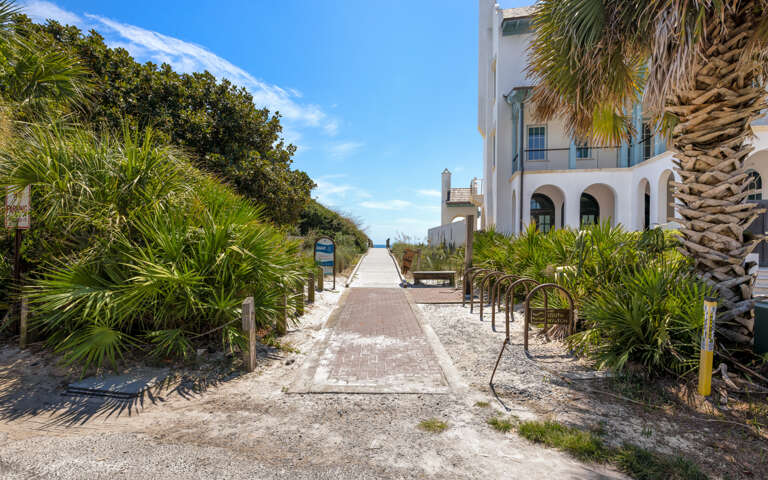 Mediterranean-style property exterior featuring a brick walkway through tropical palm landscaping leading toward beachfront access.