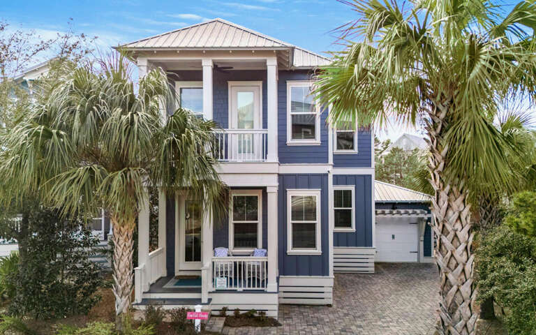 Charming coastal home with distinctive blue siding and white trim, surrounded by tropical palm trees and lush landscaping.