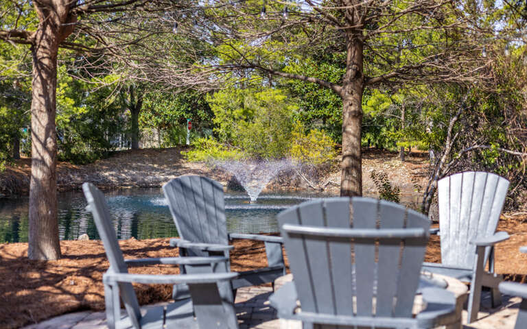 Peaceful outdoor seating area overlooking a tranquil pond with fountain, surrounded by mature trees and natural landscaping.