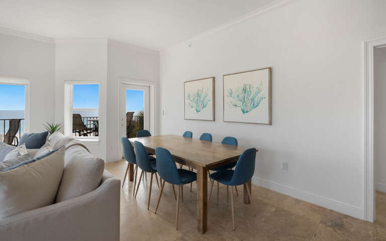 Your bright dining space opens to stunning ocean views through floor-to-ceiling windows, where coastal artwork complements the serene blue palette.