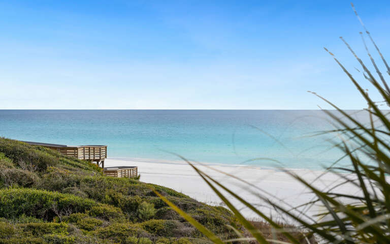 Pristine beach with crystal-clear turquoise waters and wooden boardwalk access through coastal vegetation.
