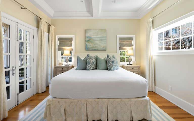 Drift into peaceful sleep on this plush bed, surrounded by warm coastal colors and soft natural light from French doors.