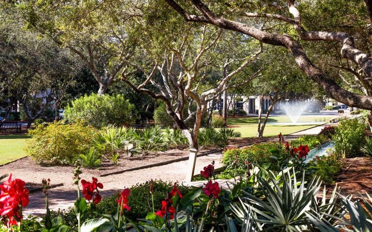 Mature trees create dappled shade across manicured gardens with colorful flowering plants and peaceful walkways throughout the property grounds.