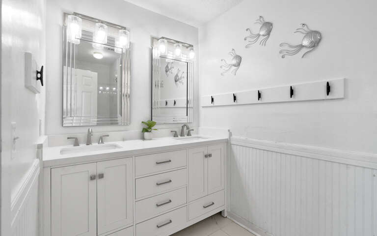 Step into your pristine white bathroom with elegant double vanities, perfect Hollywood-style lighting, and charming coastal fish art.