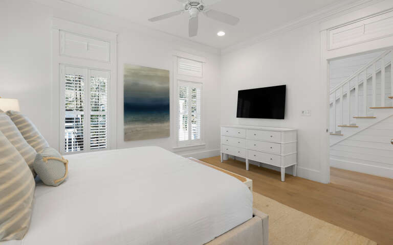 Unwind in your serene white bedroom featuring plush bedding, ocean-inspired artwork, and a wall-mounted TV for cozy nights in.