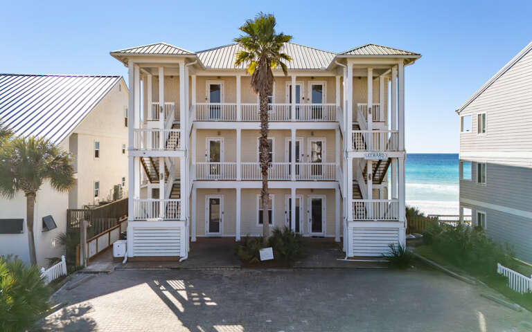 Charming gulf front building with multiple balconies overlooking turquoise waters and white sand, surrounded by swaying palms in a coastal paradise.