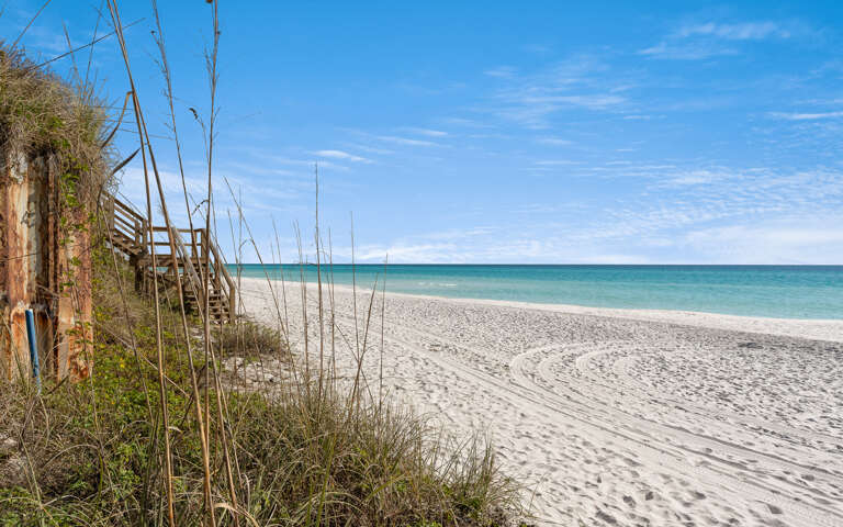 Pristine white sand beach with wooden boardwalk access through natural dunes, offering stunning turquoise waters and endless coastal views.