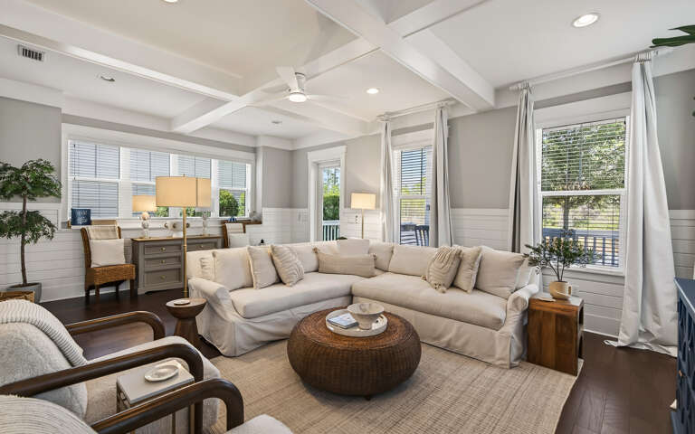 Relax in your elegant living room featuring plush sectional seating, coffered ceilings, and abundant natural light through plantation shutters.