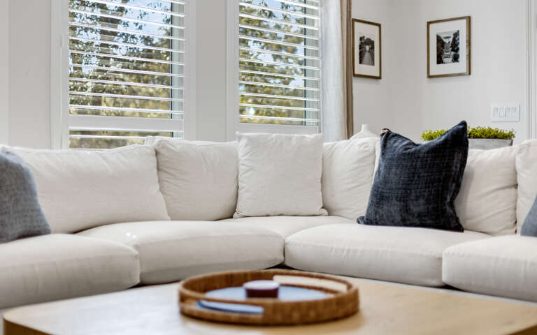 Sink into this plush white sectional sofa where natural light filters through plantation shutters, creating your perfect afternoon retreat.