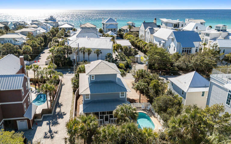 Aerial view of a charming coastal neighborhood with beach houses featuring metal roofs and private pools, just steps from pristine turquoise waters.