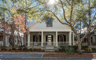 A charming white cottage with wraparound porch nestled among mature oak trees on a quiet cobblestone street.