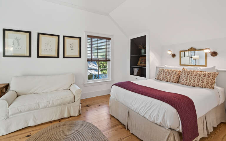 Unwind in your charming bedroom featuring a cozy reading chair and warm natural light streaming through plantation shutters onto beautiful hardwood floors.