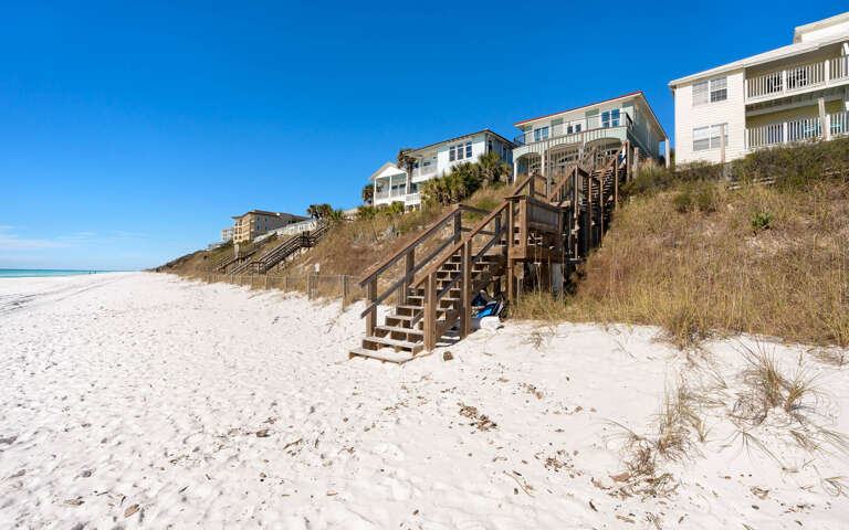 Beachfront properties with wooden staircases leading down to pristine white sand beach under clear blue skies.