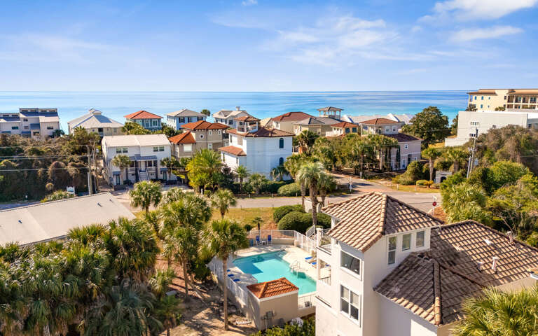 Elevated aerial view showcases a coastal neighborhood with colorful beachfront homes and palm-lined streets near pristine turquoise waters.