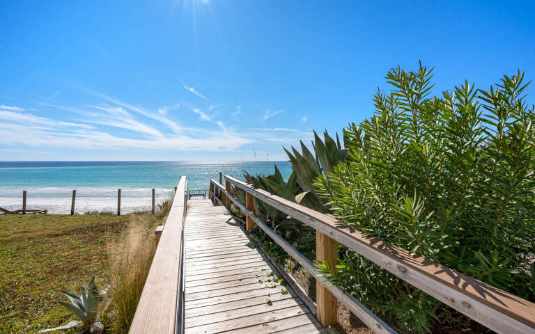 A scenic wooden boardwalk leads through coastal vegetation to pristine sandy shores and crystal-clear waters.