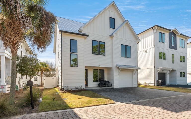 Modern coastal home featuring light-colored siding and metal roof, nestled among palm trees in a charming neighborhood setting.