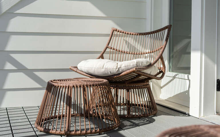 Sink into this cozy wicker chair with soft cushions, where warm sunlight creates perfect shadows for your peaceful morning coffee moments.