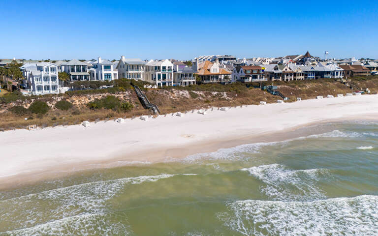 Stunning aerial view of beachfront homes nestled along pristine white sand shores with crystal-clear emerald waters.