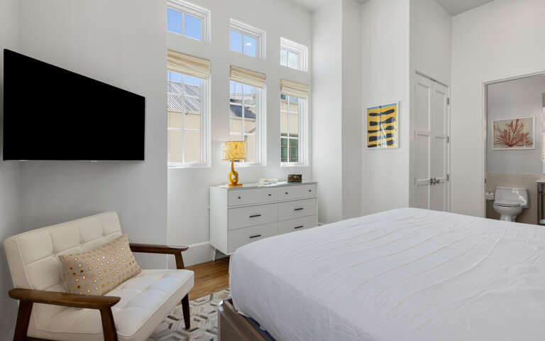 Your bright bedroom features crisp linens, a cozy reading chair, and tall windows flooding the space with natural light.