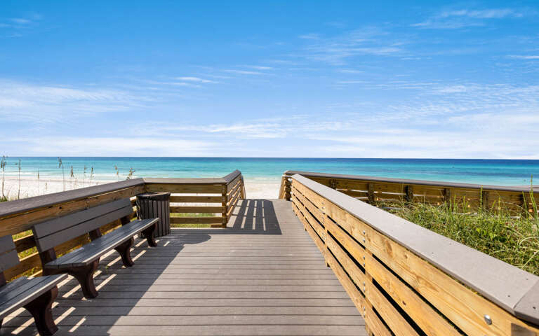 Wooden boardwalk overlooking pristine white sand beach and turquoise waters under clear blue skies.