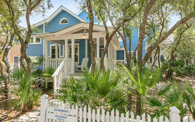 Charming blue cottage nestled among tropical palms and mature trees, featuring a welcoming front porch with classic white railings.