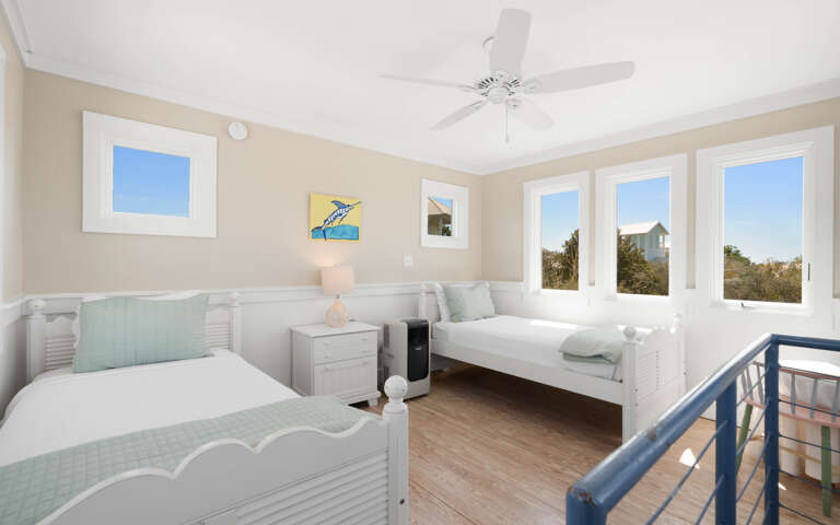Relax in this bright coastal bedroom with twin beds, ceiling fan, and stunning windows showcasing peaceful neighborhood views.