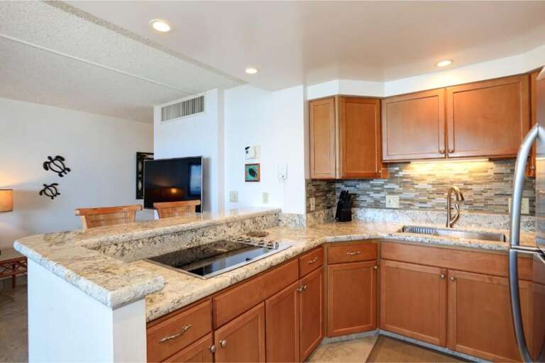 Spacious Kitchen With Stone Countertops And Stainless Steel Sink, Leading To A Living Area With Visible Television
