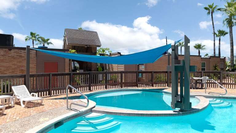 Shaded Swimming Space With Blue Sun Sail, Poolside Loungers, And A Water Feature